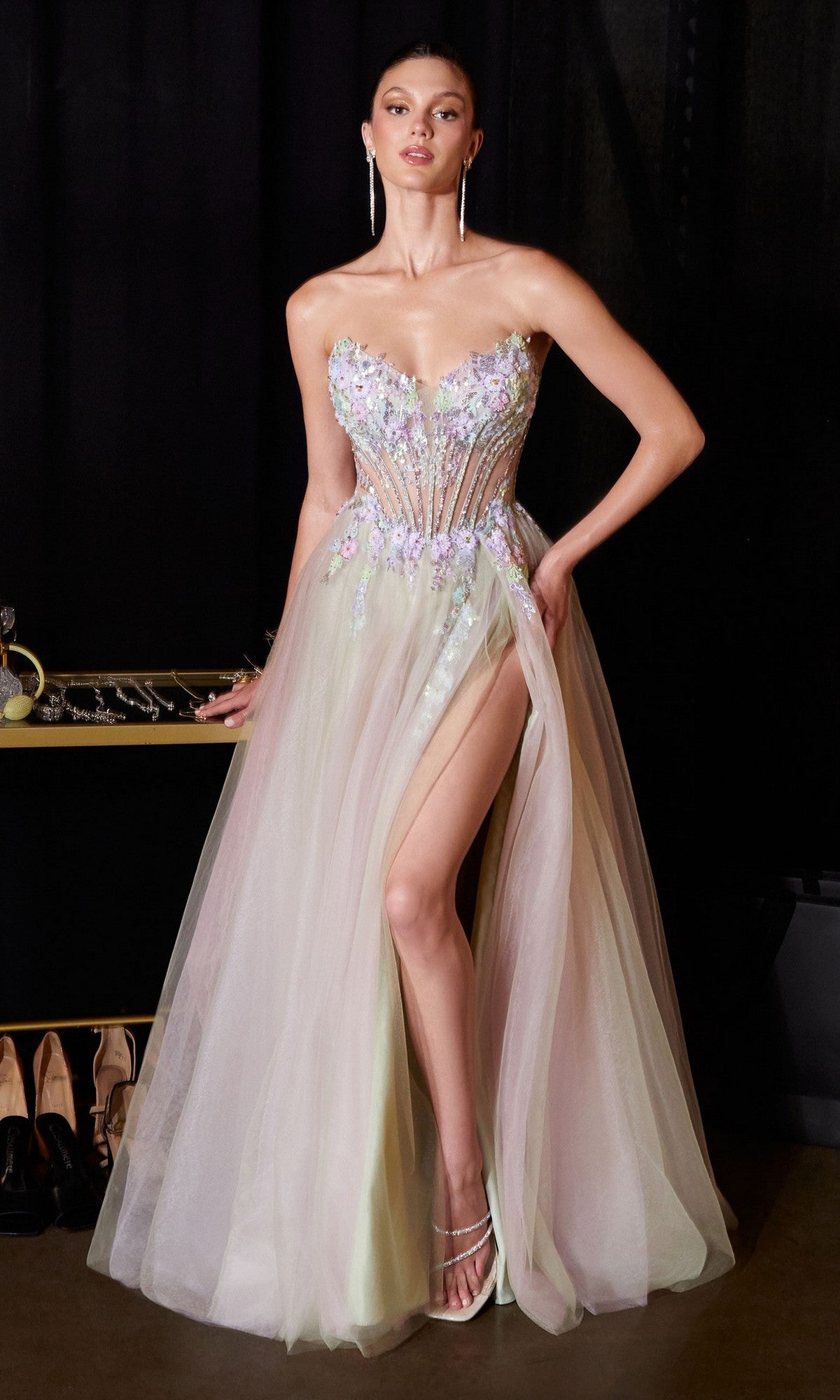 A woman wearing the Ladivine CD2418 long prom dress in sage—a strapless gown with an embroidered corset bodice, sheer tulle A-line skirt, floral adornments, and a high slit—stands with one hand on her hip.